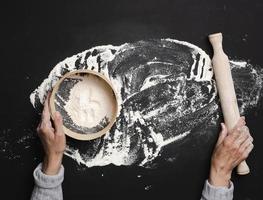 weibliche hände halten ein rundes holzsieb mit weißem weizenmehl auf einem schwarzen tisch, draufsicht foto