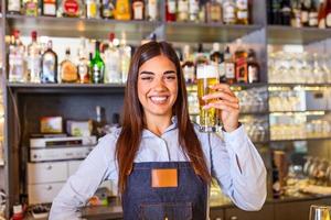 Schöne lächelnde Barkeeperin, die ein gezapftes Bier an der Theke serviert, Regale voller Flaschen mit Alkohol im Hintergrund foto