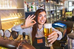 schöne lächelnde barkeeperin mit ok-zeichen, die ein gezapftes bier an der bartheke serviert, regale voller flaschen mit alkohol auf dem hintergrund foto