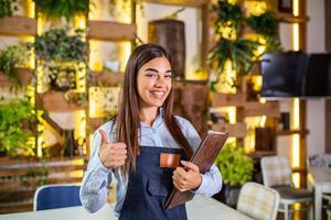 Fröhliche, schöne, lächelnde Kellnerin mit Schürze, die in einem Restaurant ein Ordnermenü gibt, in die Kamera schaut, in einem gemütlichen Kaffeehaus steht, guter Service foto