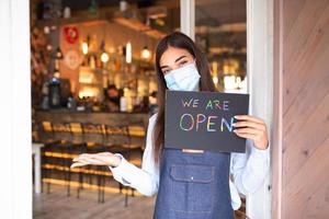 Fröhliche weibliche Kellnerin mit schützender Gesichtsmaske, die ein offenes Schild hält, während sie an der Tür eines Cafés oder Restaurants steht, wieder geöffnet, nachdem sie aufgrund des Ausbruchs von Coronavirus covid-19 gesperrt wurde foto