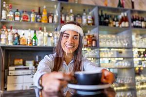 Schöne weibliche Barista hält eine Tasse mit heißem Kaffee, schaut in die Kamera und trägt einen Gesichtsschutz, während sie in der Nähe der Theke im Café steht foto