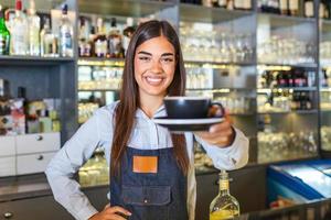 schöne weibliche barista hält eine tasse mit heißem kaffee, schaut in die kamera und lächelt, während sie in der nähe der bartheke im café steht foto