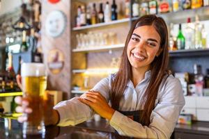 Schöne lächelnde Barkeeperin, die ein gezapftes Bier an der Theke serviert, Regale voller Flaschen mit Alkohol im Hintergrund foto