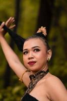 asiatische Frau, die ihr schwarzes Haar in einem grünen Kostüm hält, während sie Make-up trägt und während des Tanzfestivals vor dem Wald posiert foto