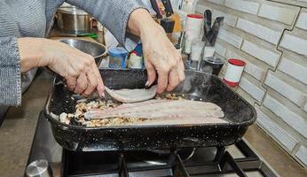 Koch legt Fisch auf Pilze mit Zwiebeln in quadratischer Pfanne auf Gasherd foto