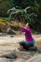 indonesische frau, die auf einem kleinen felsen sitzt und mit dem wasser spielt, während ihr kleid vom fluss nass wurde foto