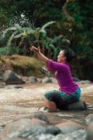 indonesische frau, die auf einem kleinen felsen sitzt und mit dem wasser spielt, während ihr kleid vom fluss nass wurde foto