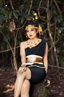 balinesische Frau sitzt wunderschön auf dem Felsen in einem schwarzen Tanzkostüm mit Make-up im Gesicht und einer goldenen Krone auf dem Kopf foto