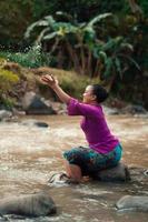 Fröhliche asiatische Frau, die mit dem Wasser in der Nähe des Flusses spielt, während sie auf dem Felsen sitzt und ein lila Kleid trägt foto