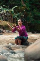 asiatische Frau, die mit schmutzigem Wasser aus einem schmutzigen Fluss spielt, während sie ein lila Kleid und einen grünen Rock trägt foto