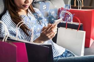 Fröhliche asiatische Frau, die auf dem Sofa sitzt und Smartphone zum Einkaufen von Online-Zahlungen und bunten Einkaufstaschen verwendet, Online-Shopping-Konzept foto
