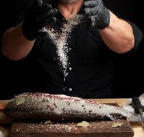 Koch in schwarzem Hemd und schwarzen Latexhandschuhen bereitet Lachsfilet auf einem Holzschneidebrett zu foto