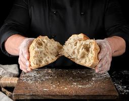 Bäcker in schwarzer Uniform brach einen halben ganzen gebackenen Laib Weißbrot aus Weizenmehl ein foto
