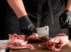 koch in schwarzen latexhandschuhen hält ein großes messer und schneidet rohes kaninchenfleisch in stücke foto