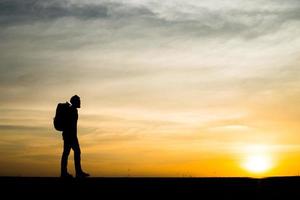 Schattenbild eines jungen Rucksacktouristenmannes, der während des Sonnenuntergangs geht foto