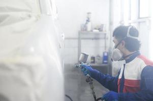 asiatischer automechaniker, der schutzanzug und atemschutzmaske trägt, lackiert karosseriestoßstangen im lackierraum und sprüht autos im lackierraum foto