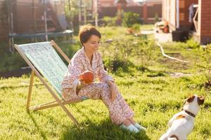 Junge Frau im Schlafanzug, die an sonnigen Sommertagen auf grünem Rasen im Stuhl ruht und Jack-Russell-Terrier-Hund - Dorf- und Landleben foto
