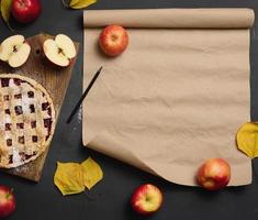 Gebackener runder traditioneller Apfelkuchen auf einem braunen Holzbrett und frischen roten Äpfeln. In der Nähe befindet sich eine braune Papierrolle zum Schreiben eines Rezepts oder Menüs, Draufsicht foto