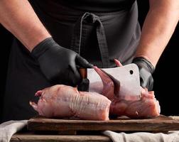 koch in schwarzen latexhandschuhen hält ein großes messer und schneidet rohes kaninchenfleisch in stücke foto