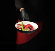Koch in schwarzer Uniform und roter Schürze hält eine runde Pfanne mit frischen Chilischoten foto