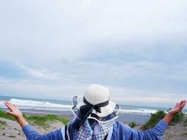 der Rücken der Frau mit Hut am tropischen Strand, die den Himmel und das Meer betrachtet, während sie ihre Hände ausbreitet foto
