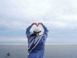 Rückseite der Frau mit Hut am tropischen Strand, die zum Himmel und zum Meer blickt, während ihre Hände Liebe formen foto