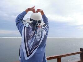 Rücken der Frau mit Hut am tropischen Strand, die zum Himmel und zum Meer aufblickte, während ihre Hände von der Brücke aus Liebe formten. Blick auf den Strand foto