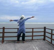 Rücken der Frau mit Hut am tropischen Strand, die den Himmel und das Meer betrachtete, während sie ihre Hände auf der Brücke ausbreitete. Seeblick foto