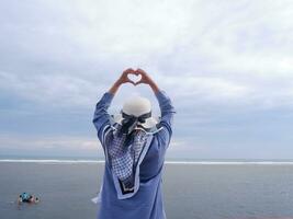 Rücken der Frau mit Hut am tropischen Strand, die zum Himmel und zum Meer blickt, während ihre Hände Liebe formen foto