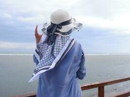 Rückseite der Frau mit Hut am tropischen Strand, die den Himmel und das Meer betrachtet, während sie ihren Hut hält foto