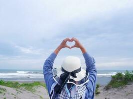 Rückseite der Frau mit Hut am tropischen Strand, die zum Himmel und zum Meer blickt, während ihre Hände Liebe formen foto