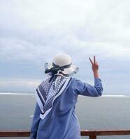 Rückseite der Frau mit Hut am tropischen Strand, die auf den Himmel und das Meer blickt und eine Friedensgeste zeigt. Vertikale. Meereshintergrund foto