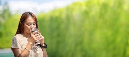 asiatische frau trinkt wasser aus einem großen glas wasser im baum-bokeh-grün im freien für bannerhintergrund. foto