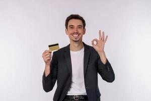 Porträt des jungen lächelnden hübschen Geschäftsmannes, der Kreditkarte und OK-Zeichenhand lokalisiert über weißem Hintergrund zeigt foto