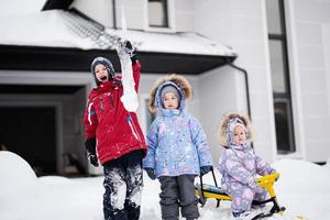 kinder spielen draußen im schnee. Drei Kinder genießen eine Schlittenfahrt. Kinderschlitten im Winter gegen Haus. foto