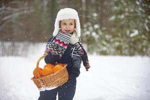 Vor dem Hintergrund eines Winterwaldes hält ein kleiner Junge mit weißer Pelzmütze einen Korb mit Orangen. Kind mit Früchten im Winter. foto