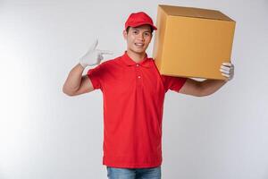 Bild eines glücklichen jungen Zustellers in roter Kappe, T-Shirt-Uniform, stehend mit leerem braunem Karton, isoliert auf hellgrauem Hintergrund Studio foto