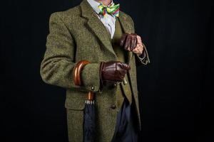 Gentleman in Tweed-Anzug und Lederhandschuhen mit Regenschirm am Arm auf schwarzem Hintergrund. Konzept des klassischen und exzentrischen britischen Gentleman-Stereotyps. Retro-Stil und Vintage-Mode. foto