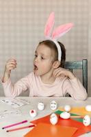 Ein kleines Mädchen in Hasenohren auf dem Kopf dekoriert weiße Eier mit Aufklebern mit verschiedenen Emotionen, in ihren Händen ist ein weißes Ei mit Emotionen, das Kind kopiert und zeigt mit Gesichtsausdrücken. foto