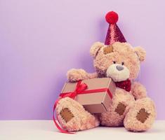 süßer brauner teddybär in einer roten kappe sitzt und hält eine braune schachtel mit einem geschenk, festlichem hintergrund foto