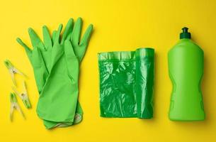 Gummigrüne Handschuhe zum Reinigen, Mülleimer-Plastikbeutelrolle und Plastikflasche mit Reinigungsmittel auf gelbem Hintergrund foto