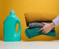 erwachsene frau faltet gewaschene trockene kleidung auf einem weißen tisch, neben einer plastikflasche mit flüssigem waschgel, gelber hintergrund foto