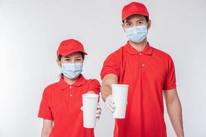 Bild eines glücklichen jungen Zustellers mit roter Kappe, leerem T-Shirt, einheitlichen Gesichtsmaskenhandschuhen, die mit leeren weißen Pappbechern isoliert auf hellgrauem Hintergrundstudio stehen foto