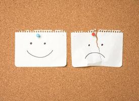 Zwei Papierblätter mit einem Lächeln und einer traurigen Emotion, die mit einem Knopf auf einem braunen Brett befestigt sind foto