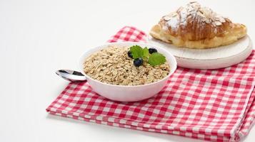 roh Haferflocken im Weiß Keramik Teller und Croissant auf Weiß Tisch, Frühstück foto