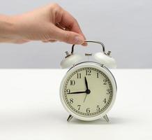 weiblich Hand hält ein Weiß Metall Alarm Uhr auf ein Weiß Tabelle foto