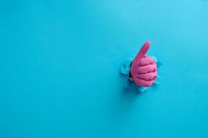 ein Hand im ein Rosa Haushalt Gummi Handschuh Stöcke aus von das zerrissen Loch und zeigt an ein Geste Okay, Daumen hoch. Blau Hintergrund foto