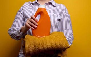 ein Frau im ein Weiß gestreift Hemd hält ein Orange Plastik Flasche mit Flüssigkeit Waschen Gel und ein gestrickt Pullover. Wäsche und Haushalt Hausarbeiten, sauber gewaschen Artikel foto