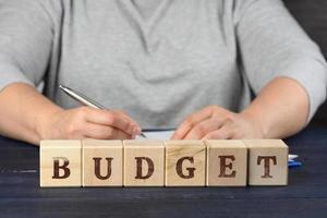 Frau sitzt beim das Tabelle und Zeichen Unterlagen mit ein Stift, im Vorderseite von hölzern Würfel mit das Inschrift Budget foto